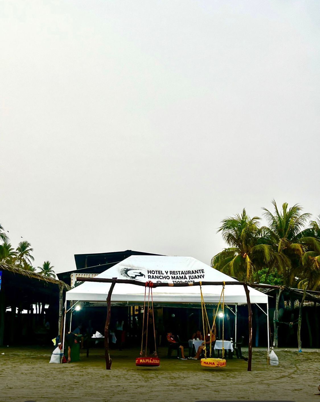 Una carpa blanca con un letrero que dice 'Hotel y Restaurante Rancho Mamá Juan' sobre arena, con palmeras.