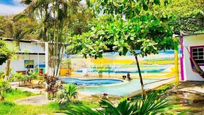 Un área de piscina con un mural, rodeada de follaje y un edificio con un ventanal morado.