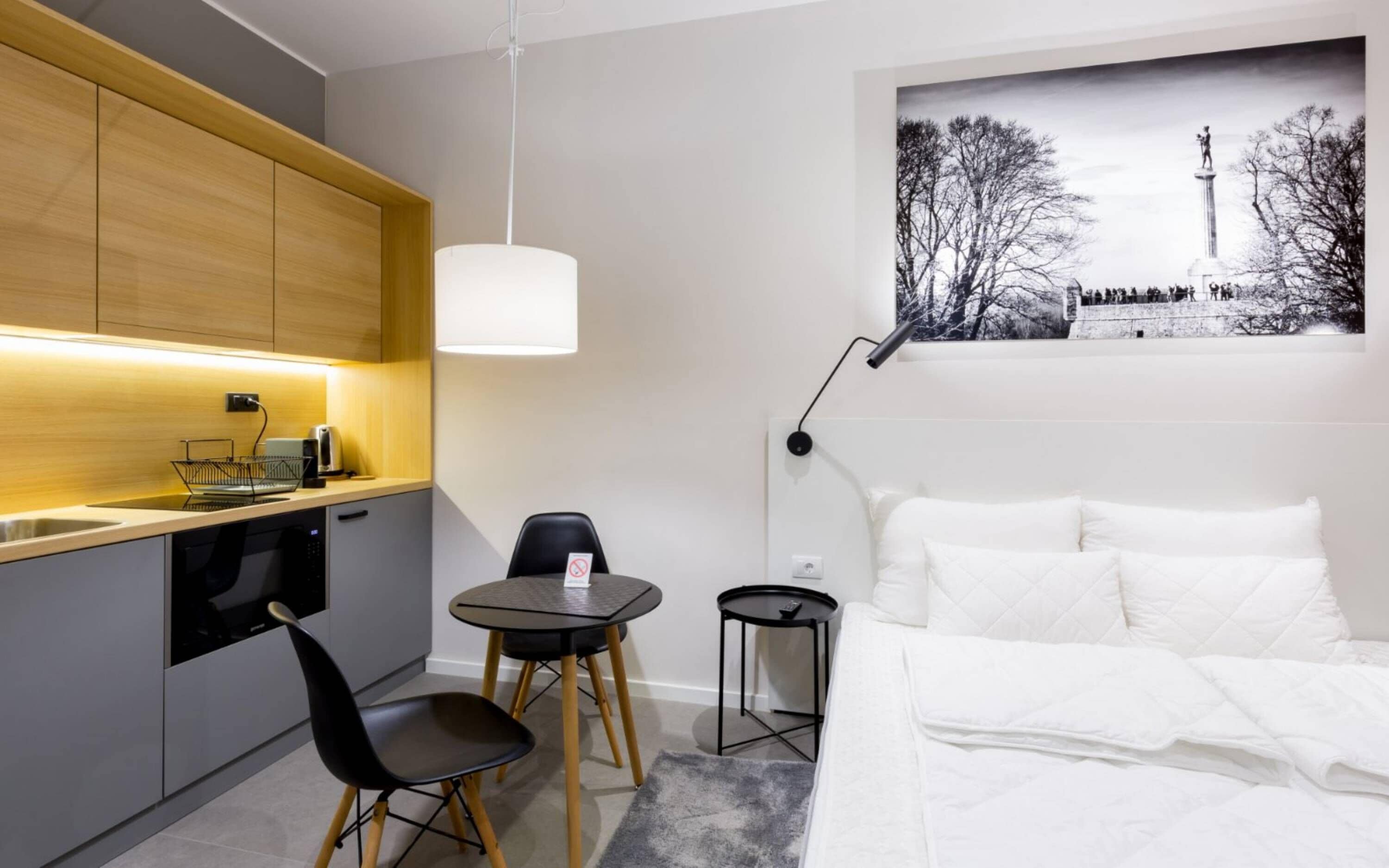 A compact hotel room with a kitchenette, a bed with white linens, a small dining table, and a chair.