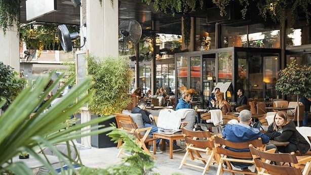 An outdoor cafe with wooden chairs and tables, surrounded by greenery and modern architecture. - Teanna Lux Apartments (Belgrade)