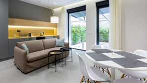 A modern living room with a sofa, coffee table, and dining table with chairs. There is a kitchen area with a countertop and appliances. Large windows provide natural light and views of greenery outside. - Teanna Lux Apartments (Belgrade)