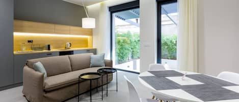 Una sala moderna con sofá, mesa de centro y mesa de comedor con sillas. Cuenta con una zona de cocina con mostrador y electrodomésticos. Grandes ventanales ofrecen luz natural y vistas al verdor exterior.