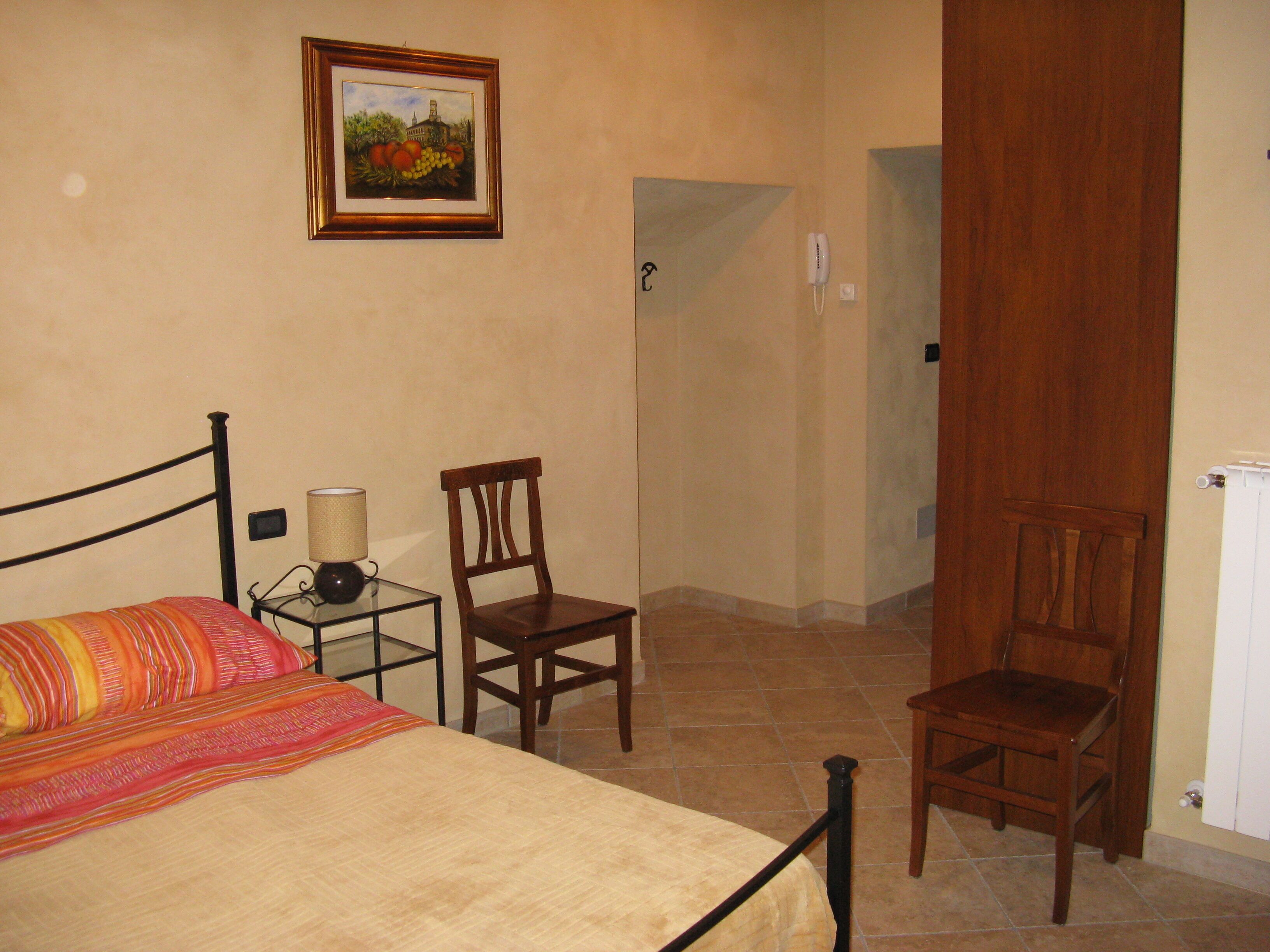 Una camera da letto con un letto, due sedie, un comodino con una lampada, un quadro appeso al muro e una porta che dà su un'altra stanza.