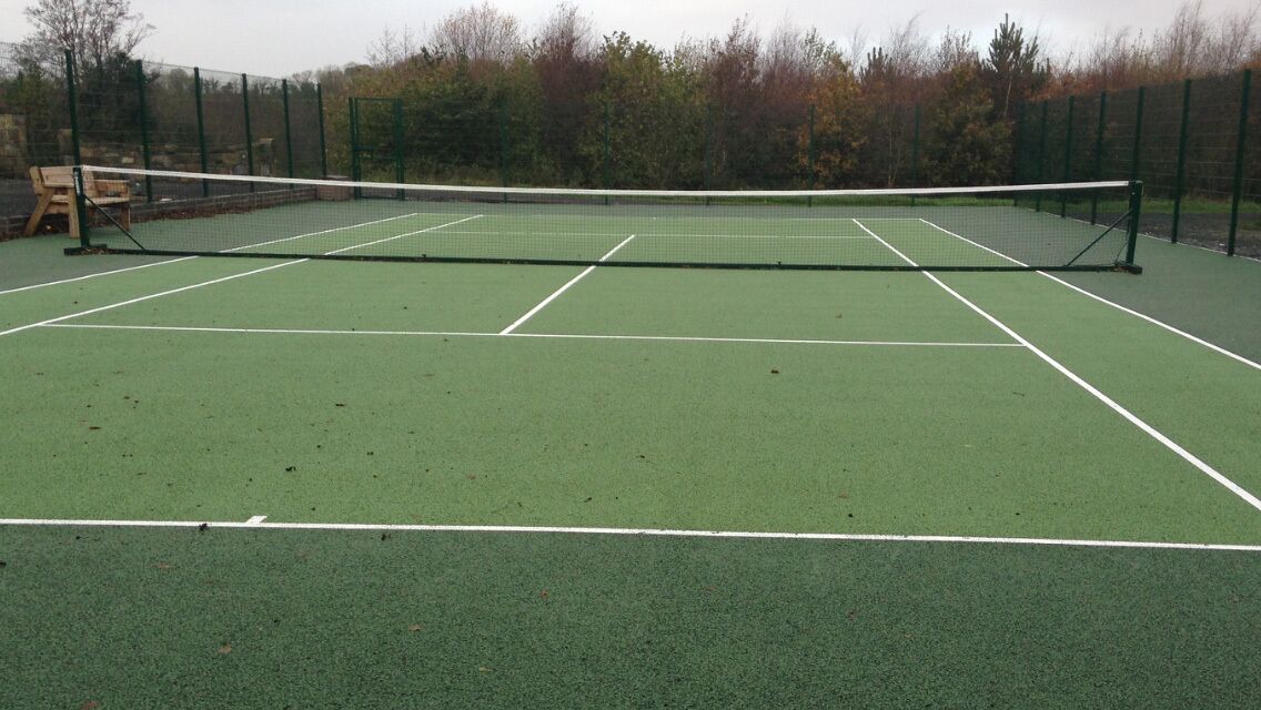 A tennis court with a net, surrounded by a green fence and trees in the background.