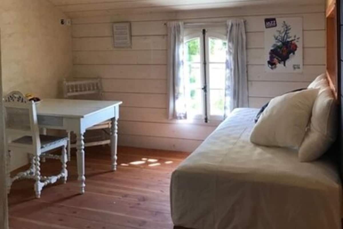 Una habitación con piso de madera, una cama con ropa blanca, una mesita y una silla. Se ve una ventana con cortinas y un cartel en la pared.