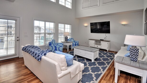 A spacious living room with a high ceiling, large windows, a ceiling fan, a flat-screen TV, and a patterned rug. - Sea Loves Me Knot - Private Pool, Elevator, Tiki Bar, 20 second walk to beach (Ocean Isle Beach)