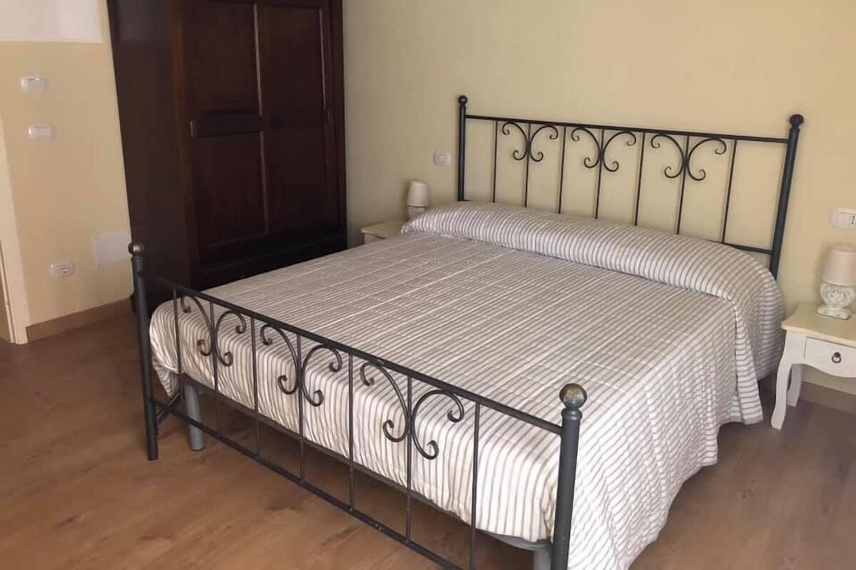 Un lit simple avec un cadre en métal et une couvre-lit à rayures, une armoire en bois et une table de chevet avec une lampe.