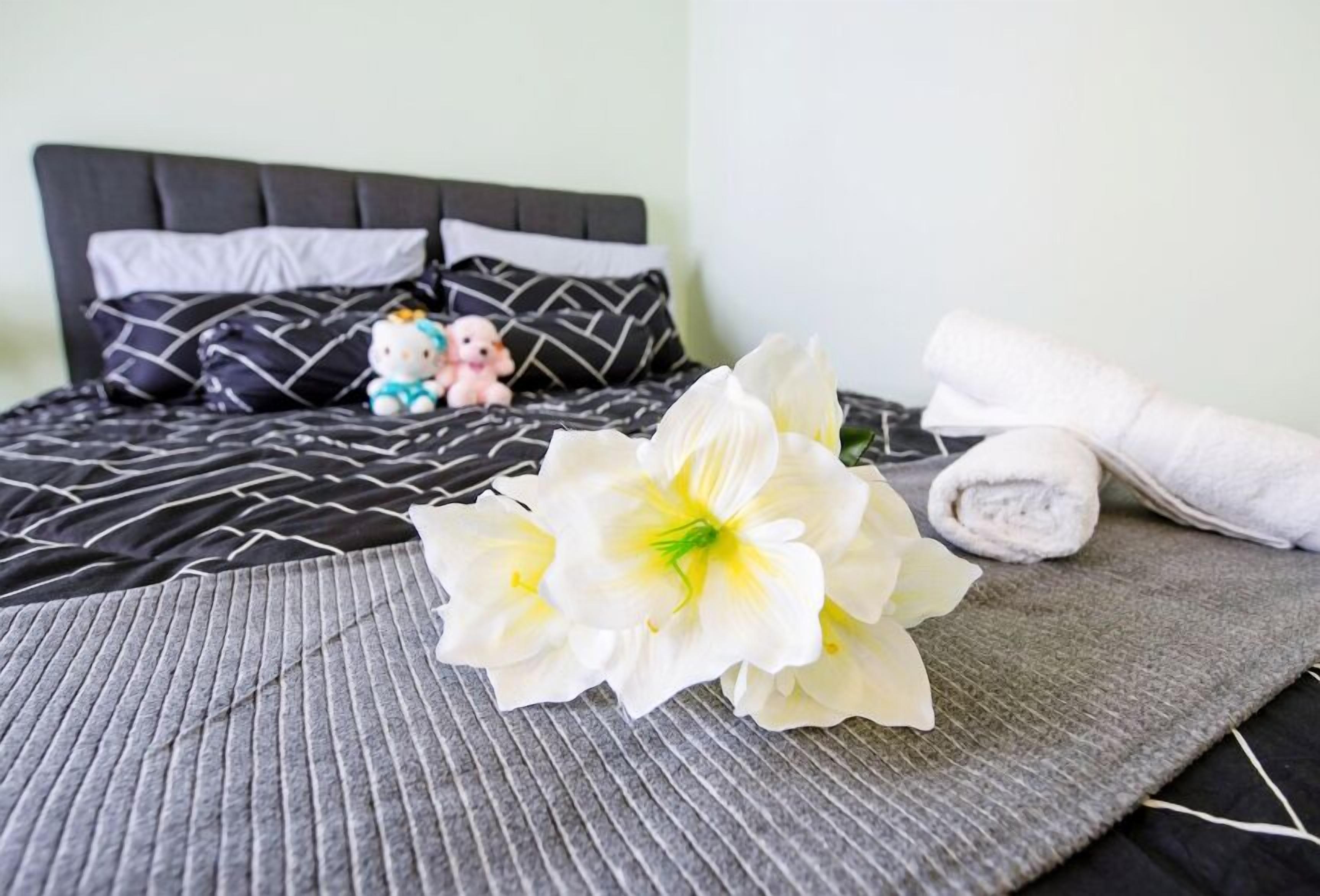 Uma cama bem arrumada com um edredom preto e branco estampado, um brinquedo de pelúcia Hello Kitty, uma figurinha Hello Kitty e uma toalha branca enrolada em um tapete cinza texturizado.