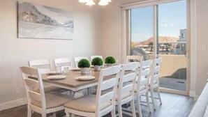 A dining area with a table set for four, white chairs, two potted plants, and a painting on the wall.