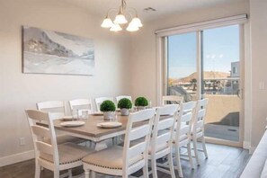 A dining area with a table set for four, white chairs, two potted plants, and a painting on the wall.