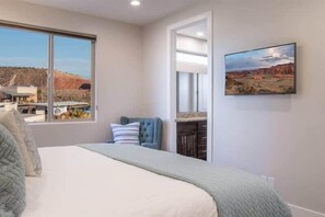 Un dormitorio con una cama grande, una ventana con vista a un edificio y paisaje, una silla y un cuadro en la pared.