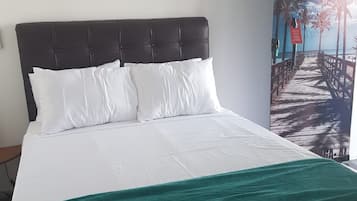 Quarto de hotel com cama, roupa de cama branca, manta azul-esverdeada e uma grande pintura de uma cena de praia.