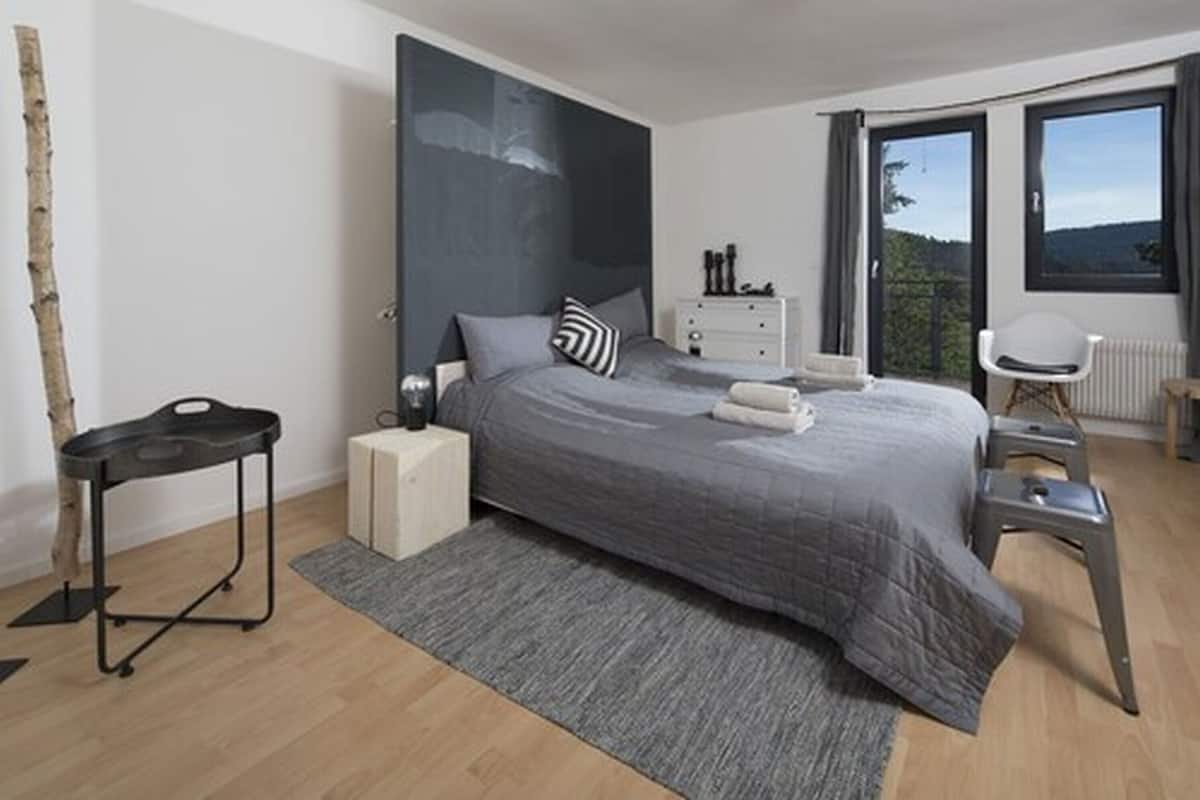 Une chambre à coucher moderne avec un grand lit, une commode, un fauteuil et une vue sur l’extérieur.