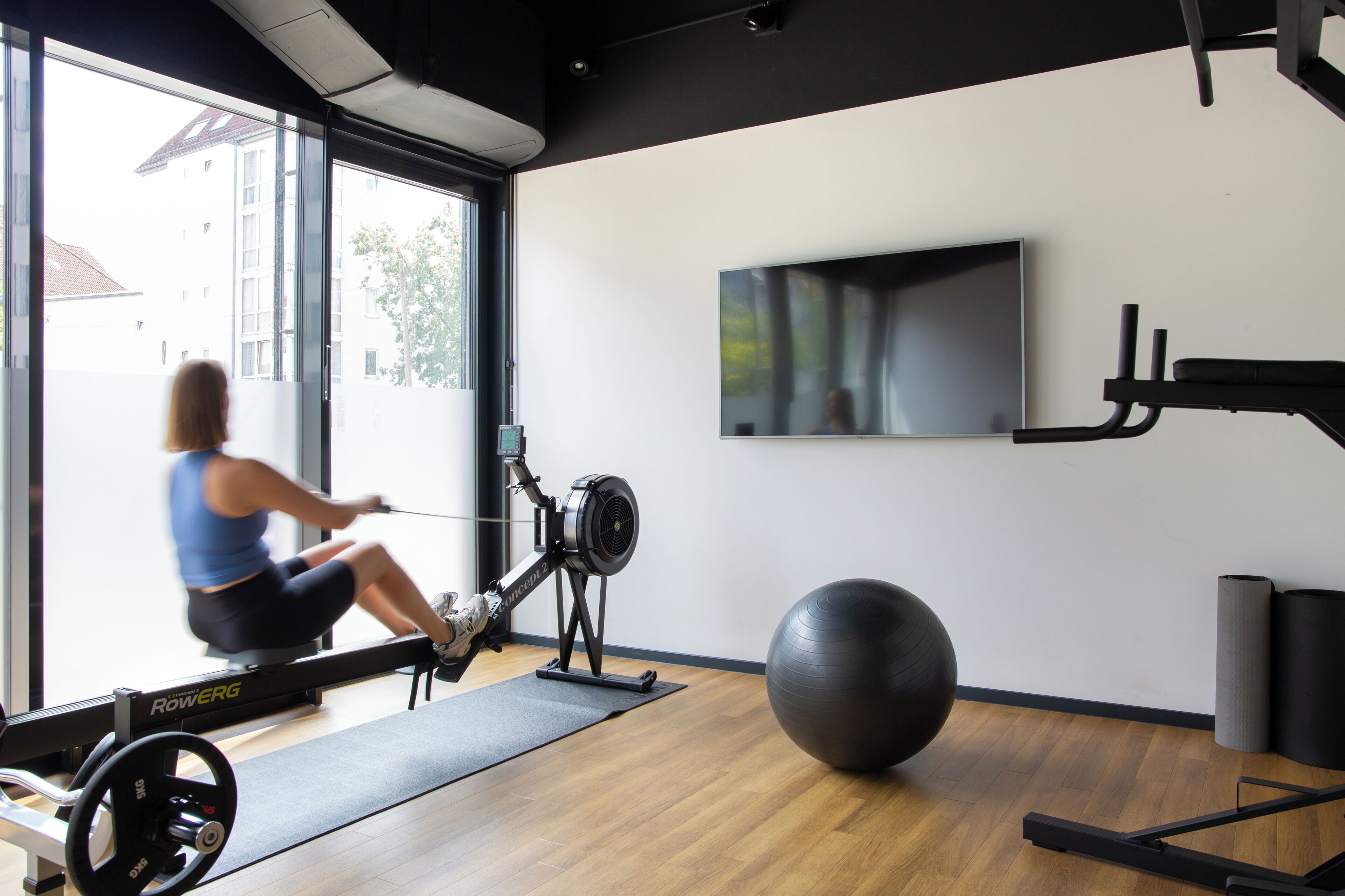 A gym with a rowing machine, exercise ball, and a TV on the wall.