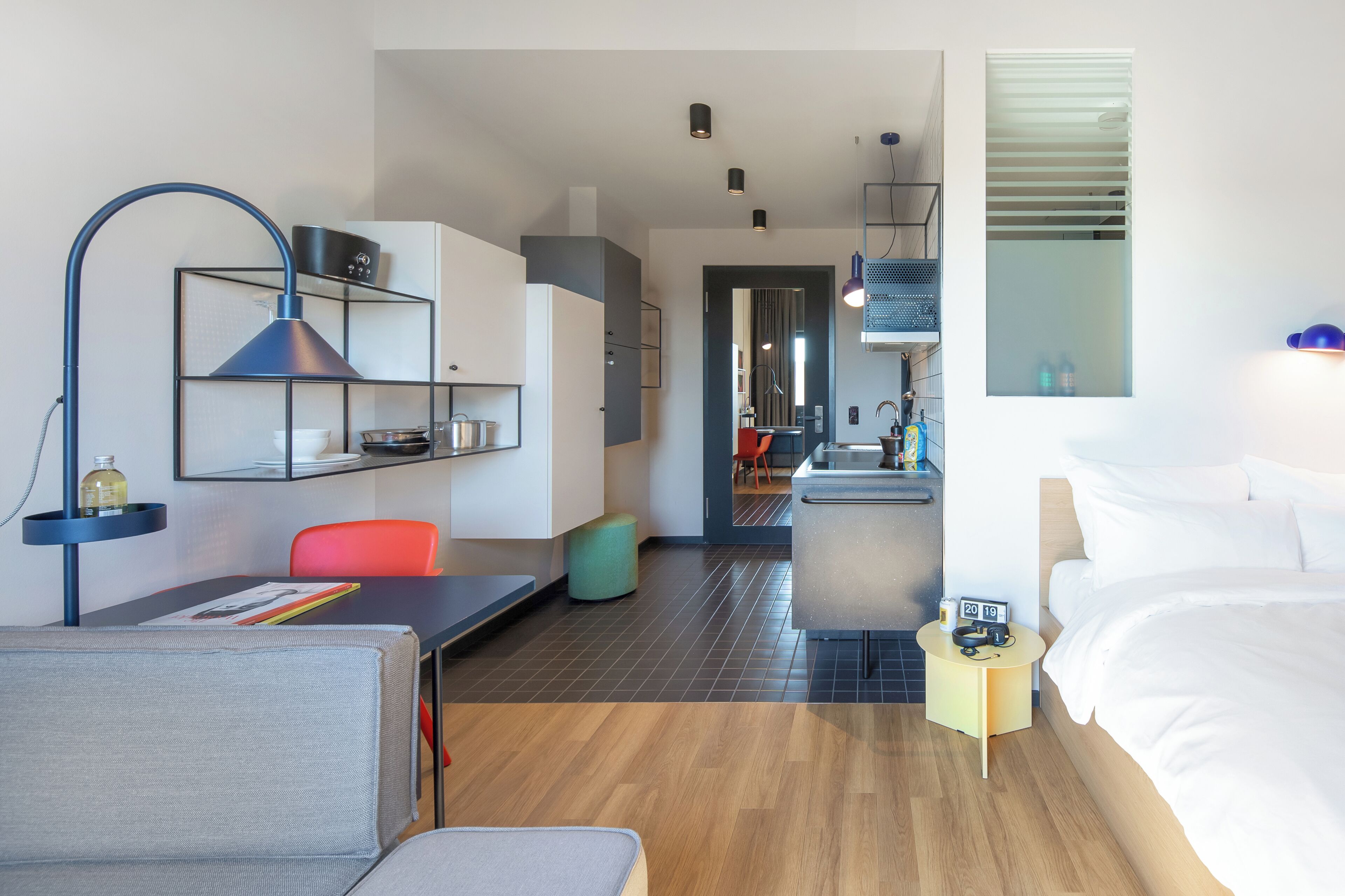 A modern hotel room with a bed, a desk, a chair, a small kitchen area, and a bathroom visible through an open door.