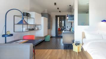 A modern hotel room with a bed, a desk, a chair, a small kitchen area, and a bathroom visible through an open door.