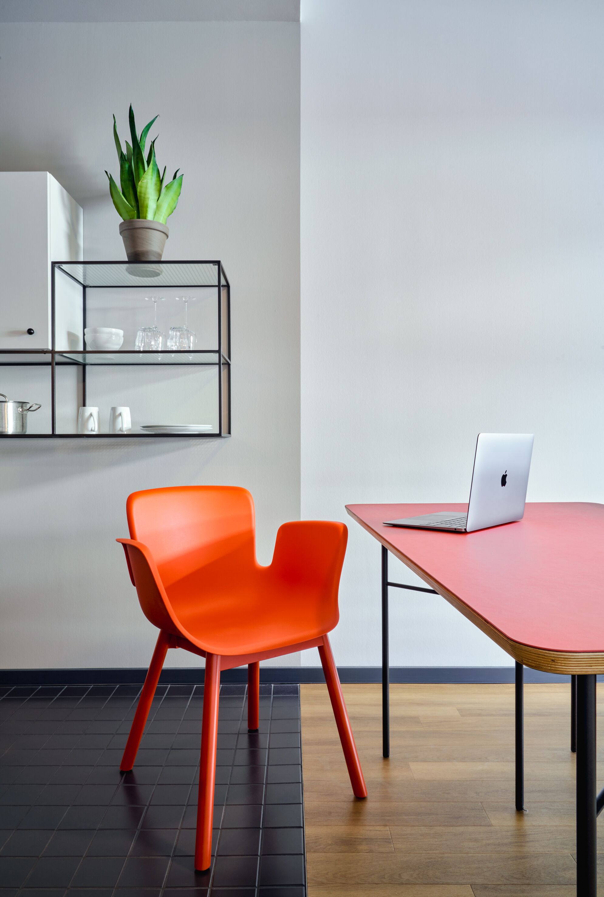 A modern room with a red table, a laptop, a bright orange chair, a potted plant, and a glass shelf with kitchen items.