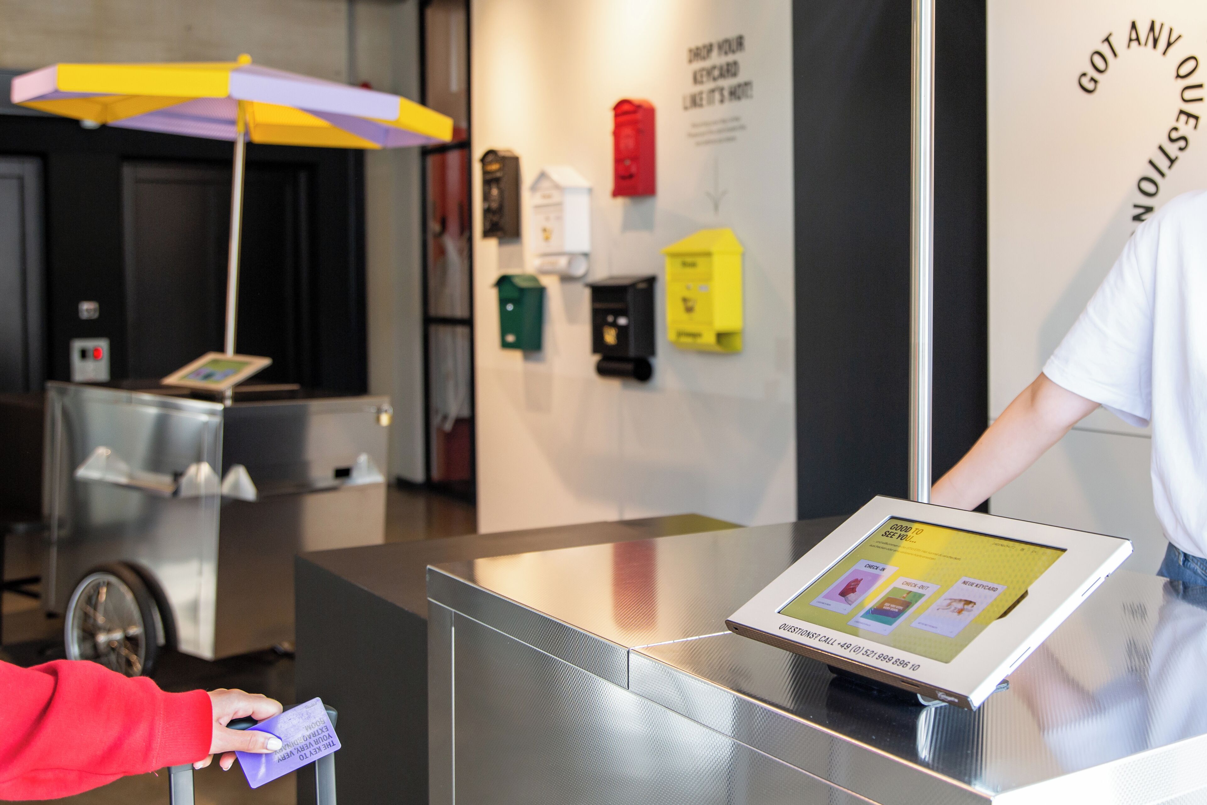 A self-service kiosk with a touchscreen interface, a person holding a card, and a vending machine in the background.