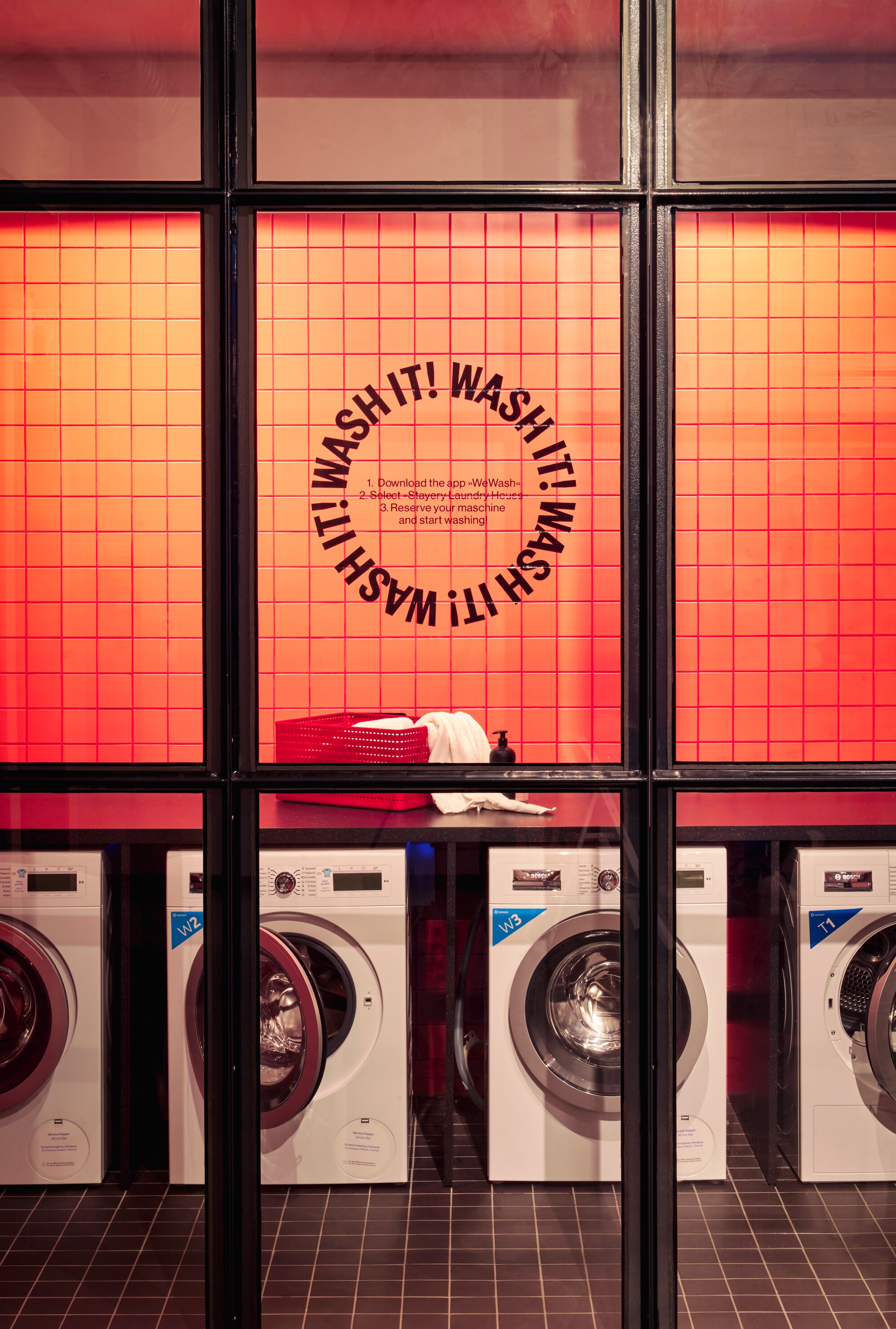 A laundromat with multiple washing machines and dryers, a red tiled wall with a circular text design, and a glass partition.