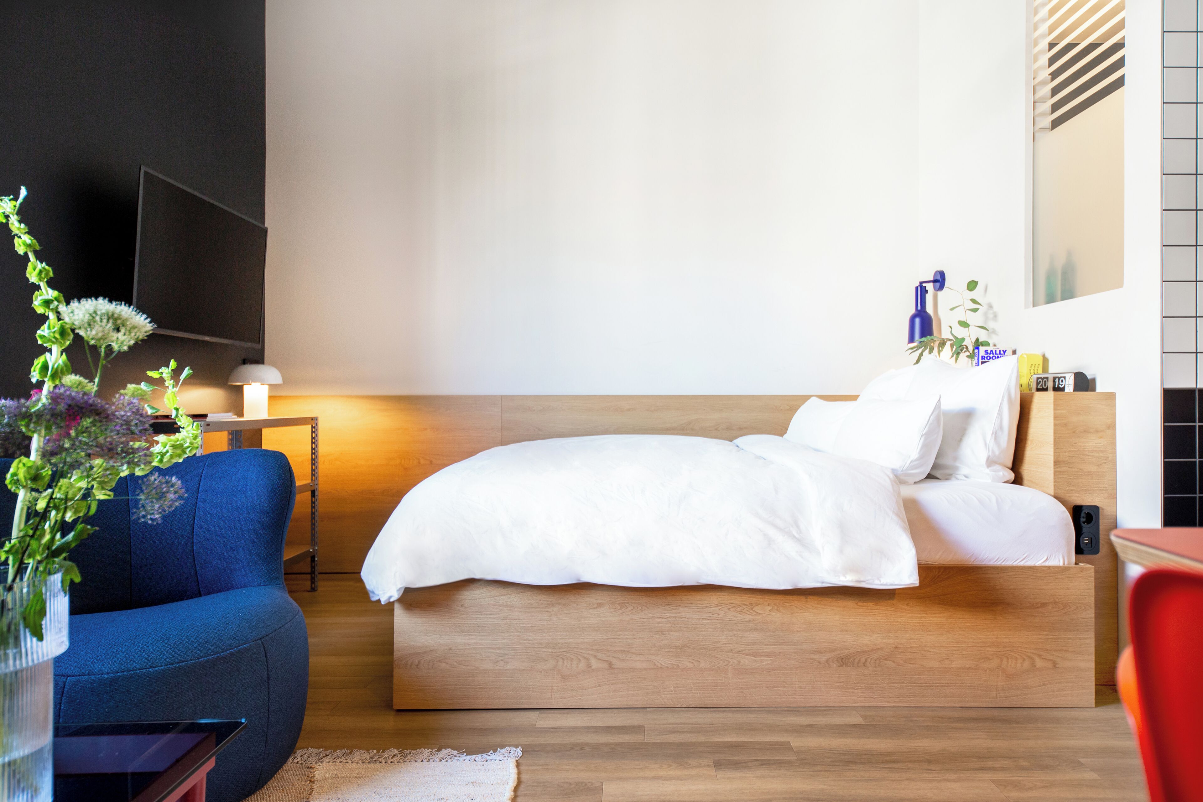 A modern hotel room with a wooden bed, a blue armchair, a small table with a lamp, and a TV mounted on the wall.