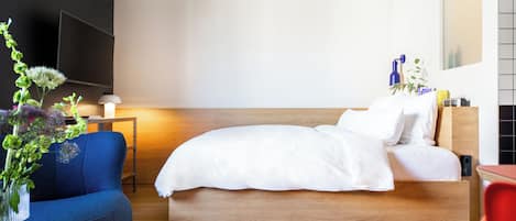 Habitación de hotel moderna con una cama de madera, un sillón azul, una mesita con una lámpara y un televisor instalado en la pared.