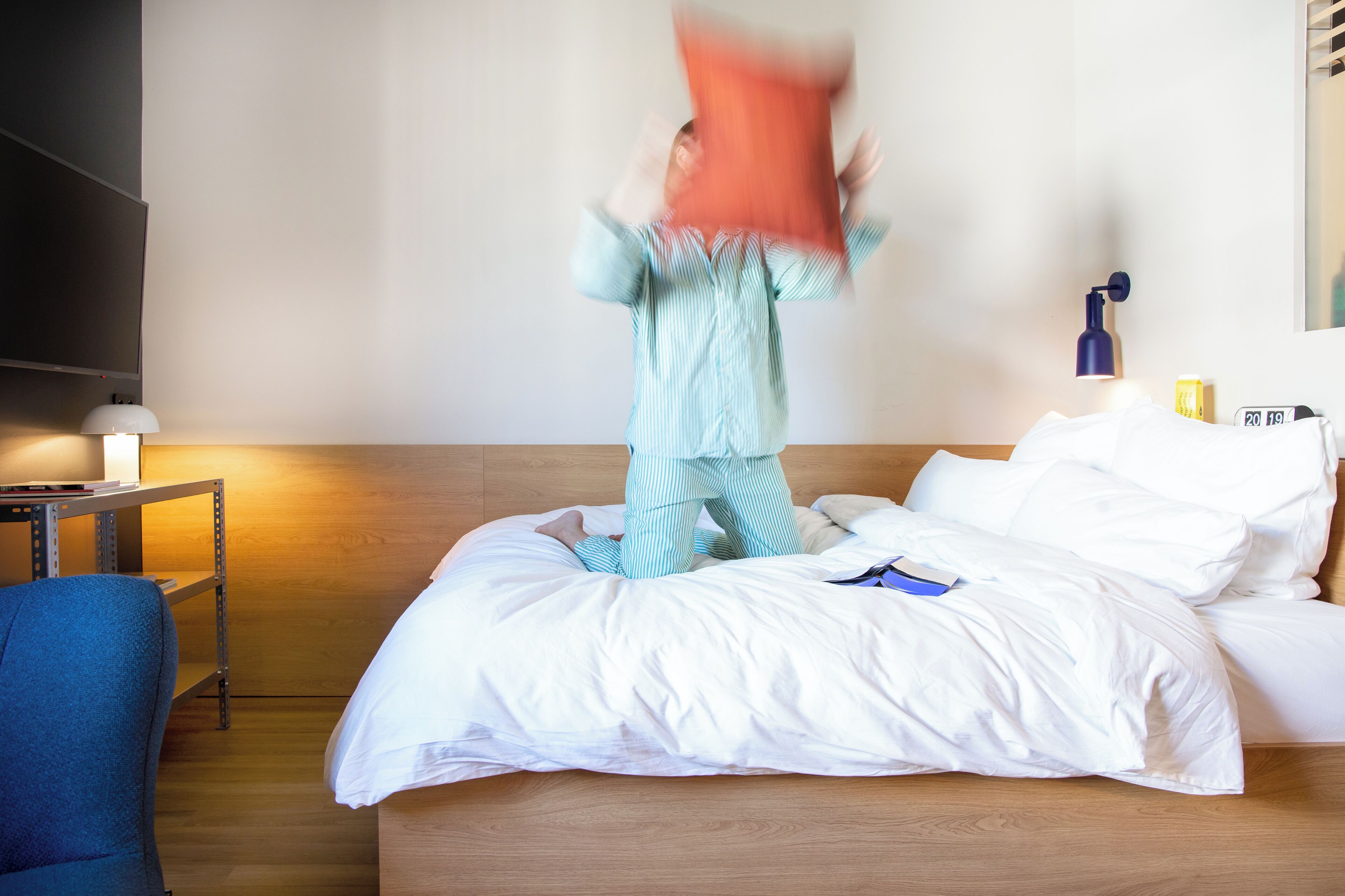 Quarto de hotel com uma cama, uma mesa de cabeceira com abajur, uma cadeira azul e uma prateleira na parede.