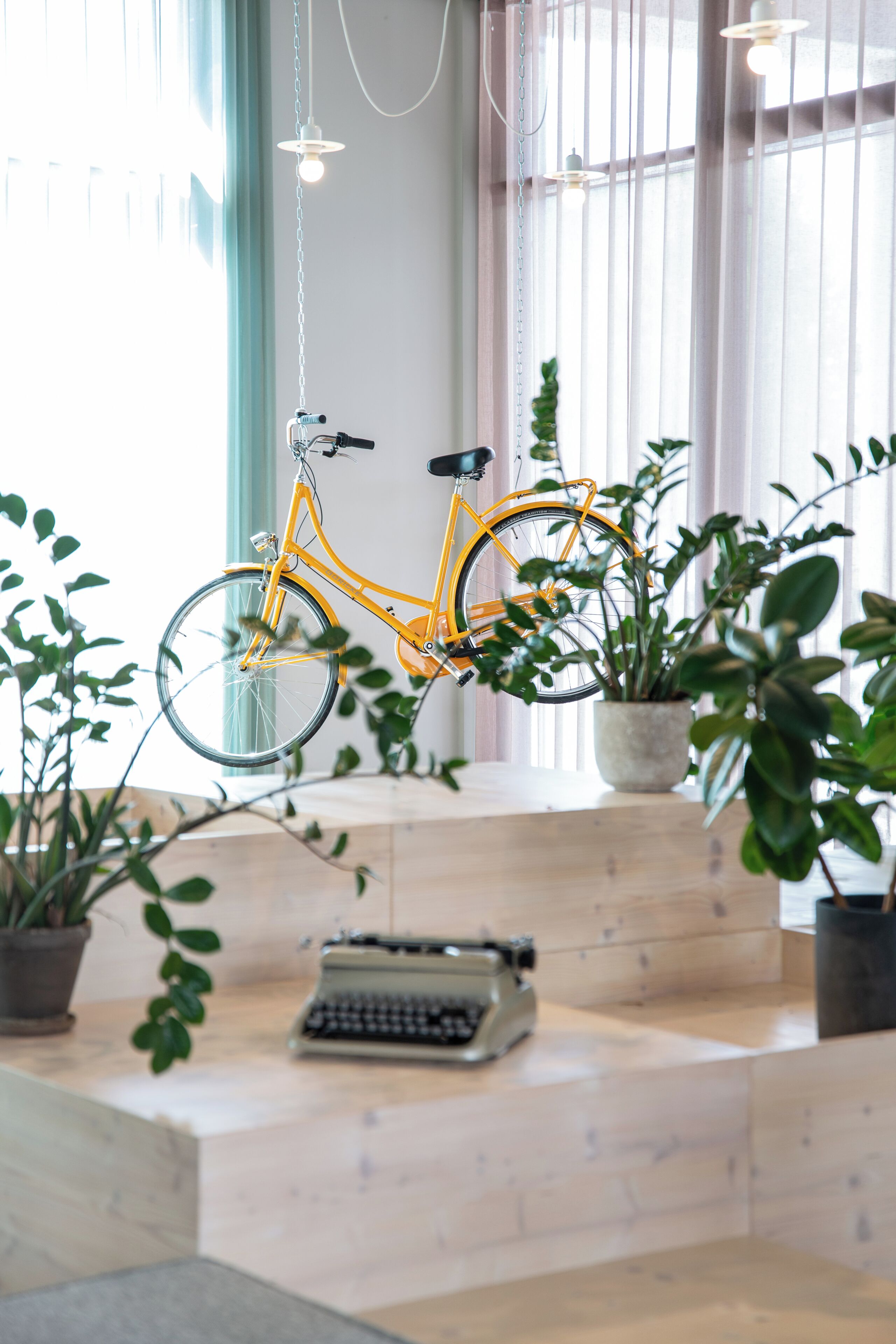 Una bicicleta amarilla colgada en una pared, una máquina de escribir y plantas en macetas en una habitación con ventanas amplias.
