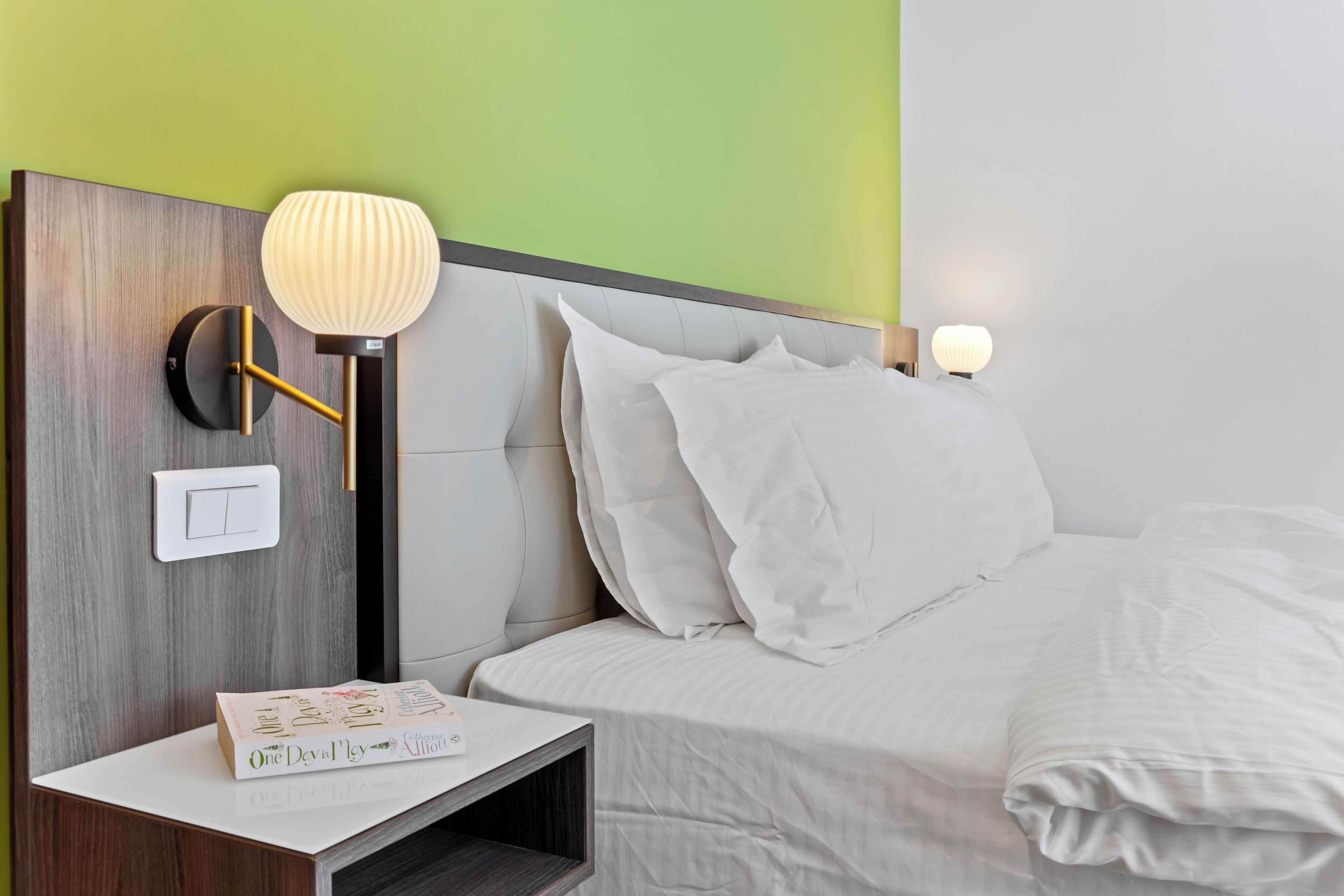A neatly made bed with white bedding, a bedside table with a book, and a wall-mounted lamp.