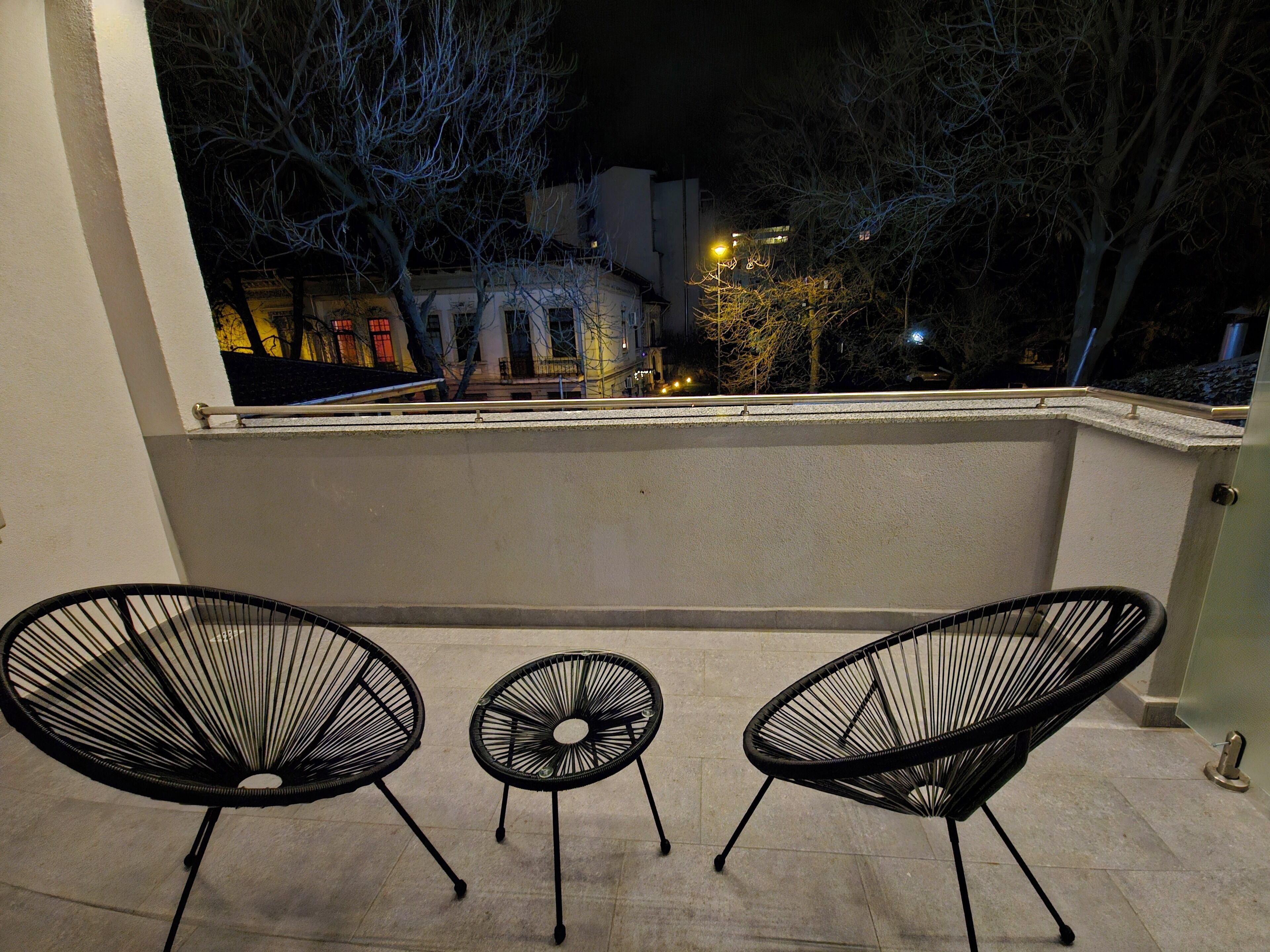 A balcony with three black wire-framed chairs, a small round table, and a view of a cityscape at night.