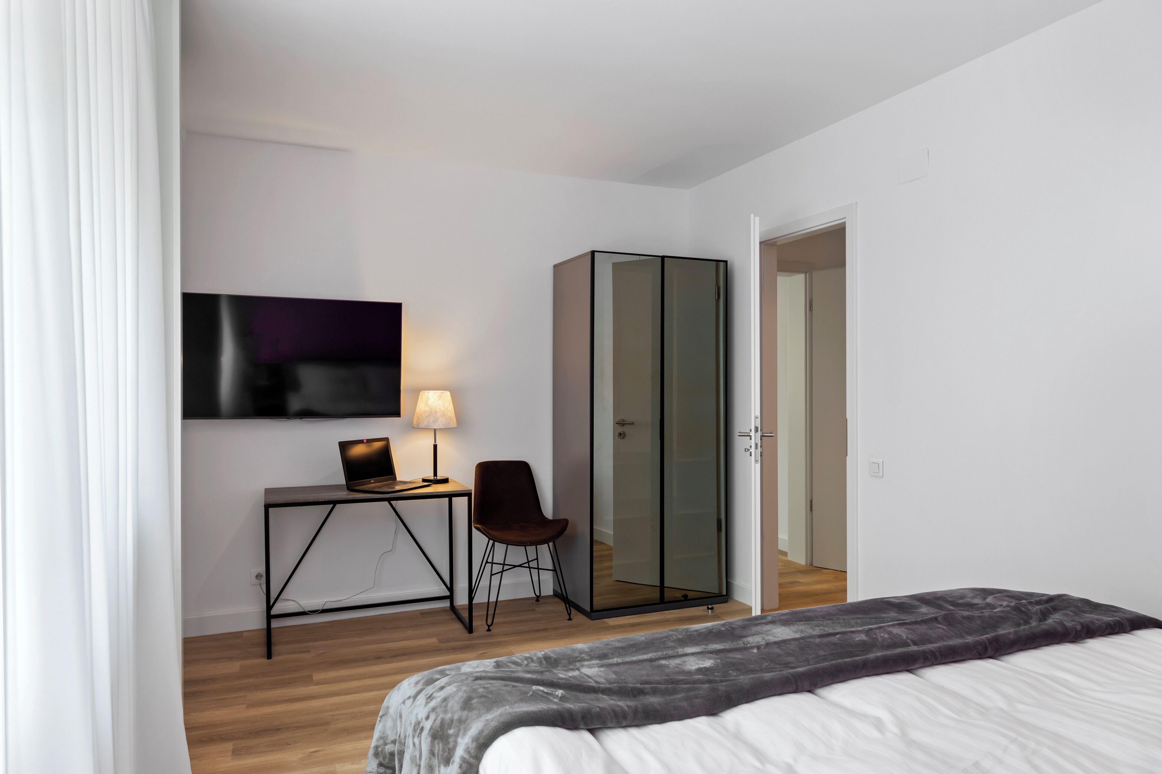 A modern hotel room with a bed, a desk with a laptop, a chair, a TV, and a wardrobe.