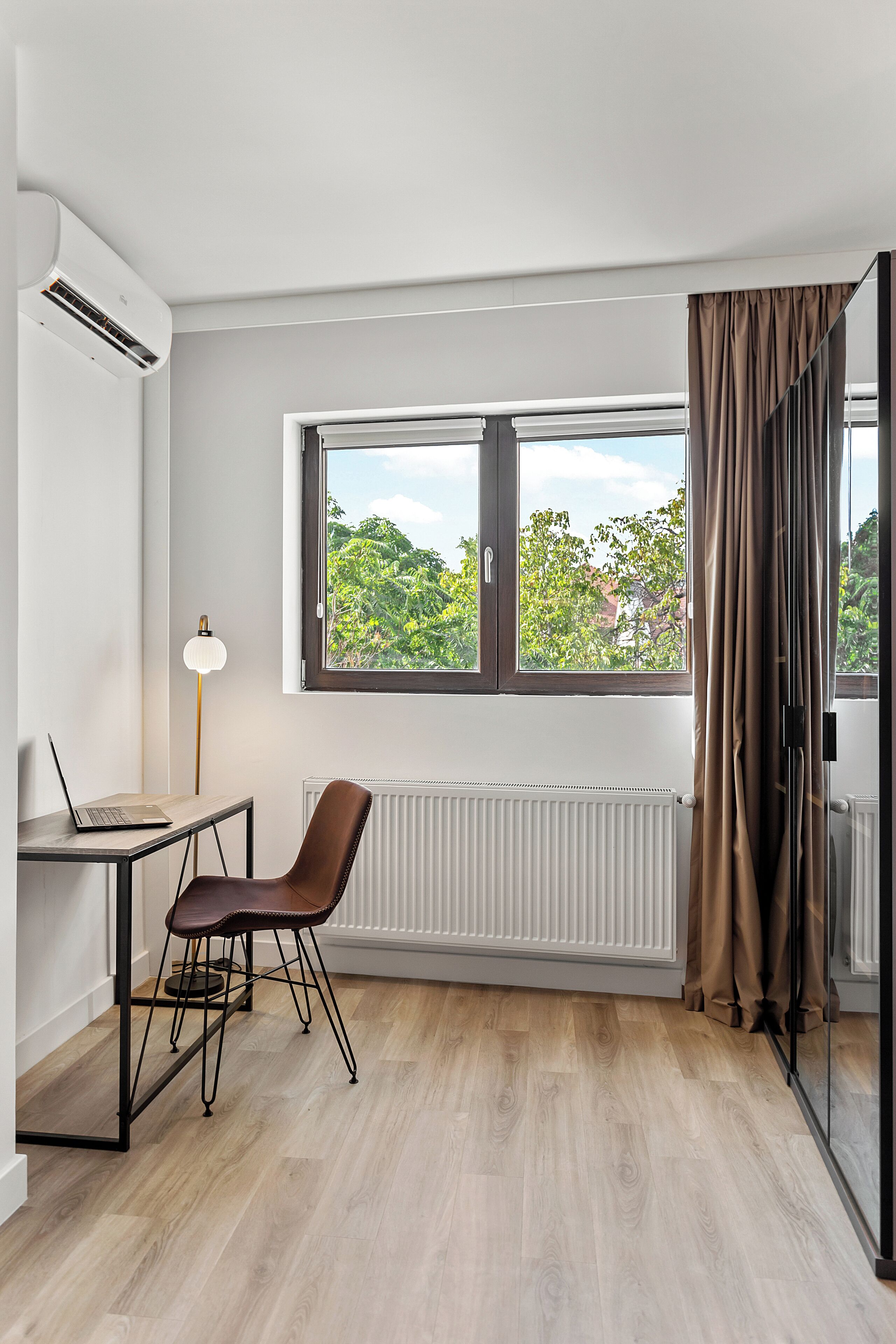 A modern room with a wooden floor, a desk with a laptop, a chair, a radiator, and a window with curtains.