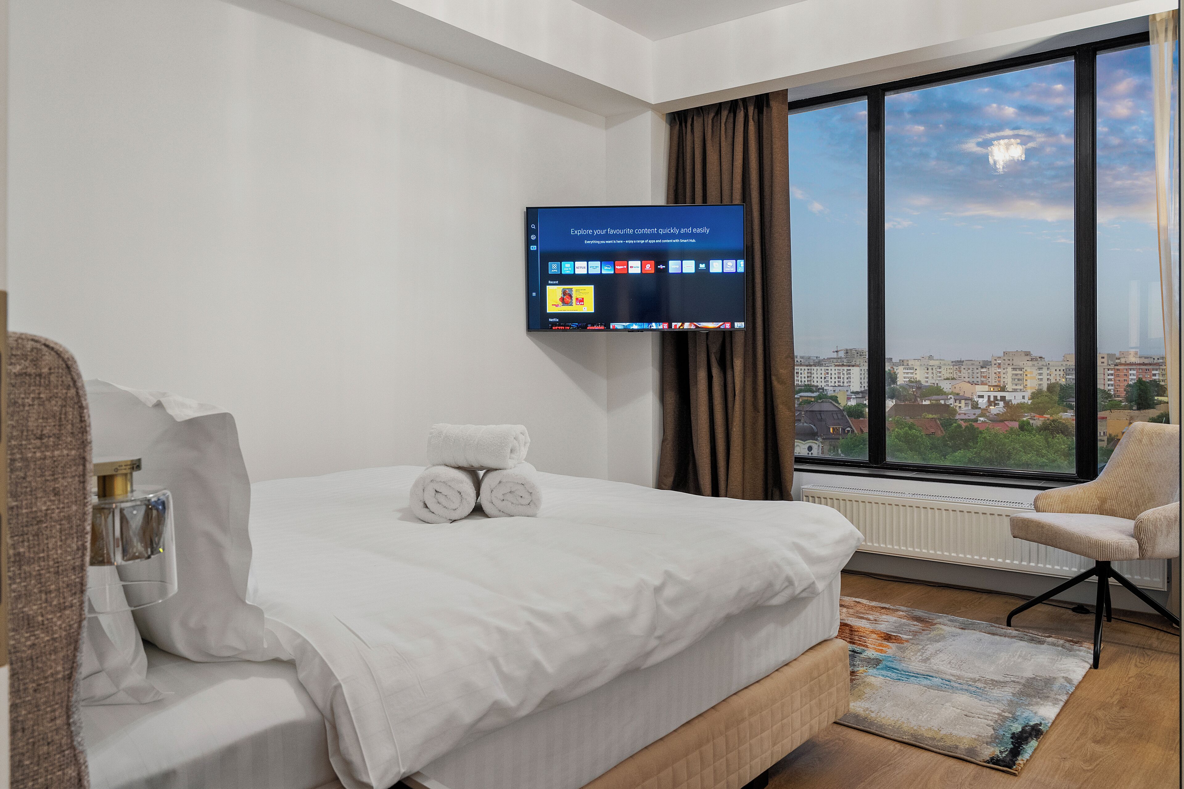 Habitación de hotel con un ventanal grande, cama con ropa blanca, televisor de pantalla plana montado en la pared y una silla junto a la ventana.