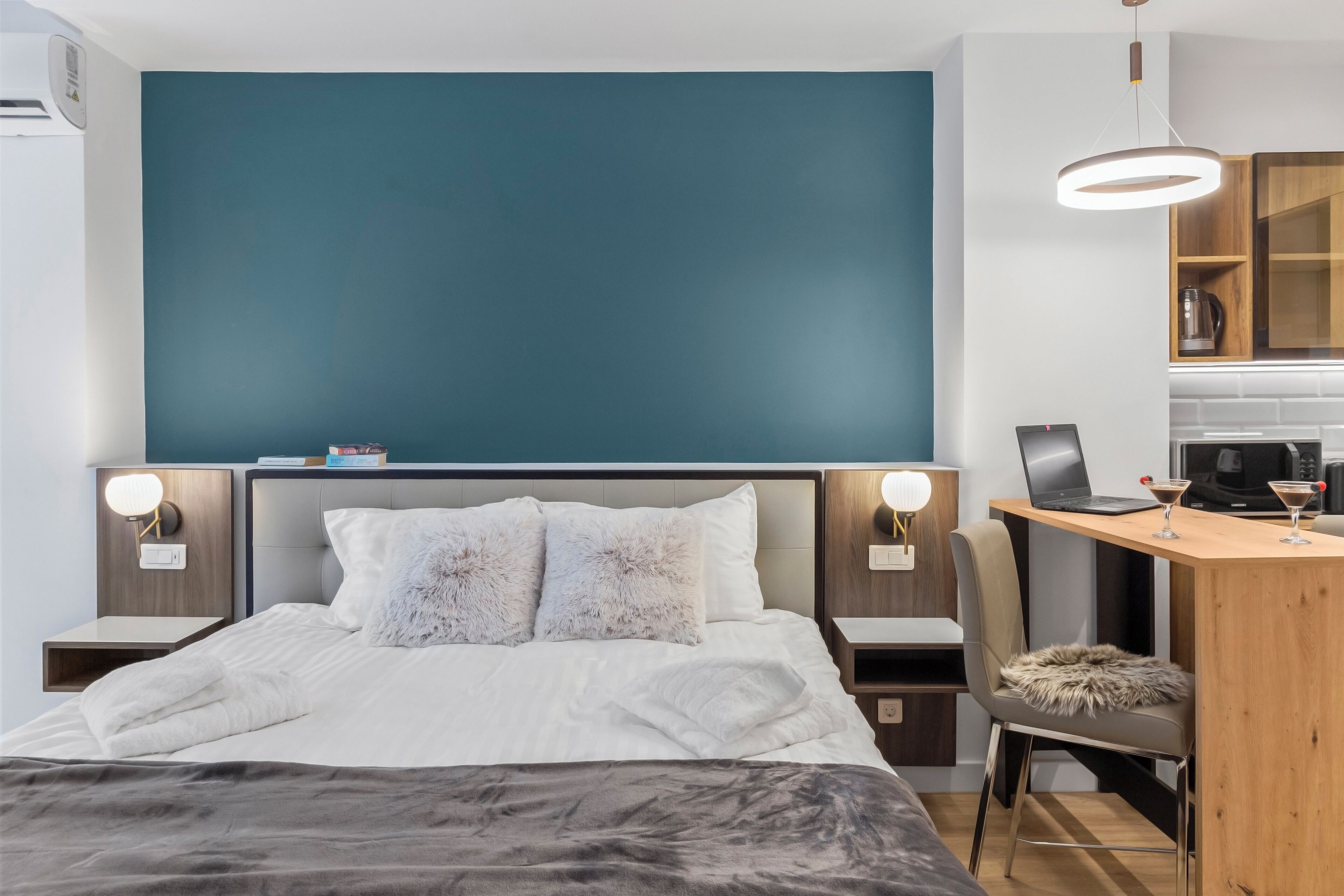 A modern bedroom with a bed, bedside tables, a desk, and a kitchenette.