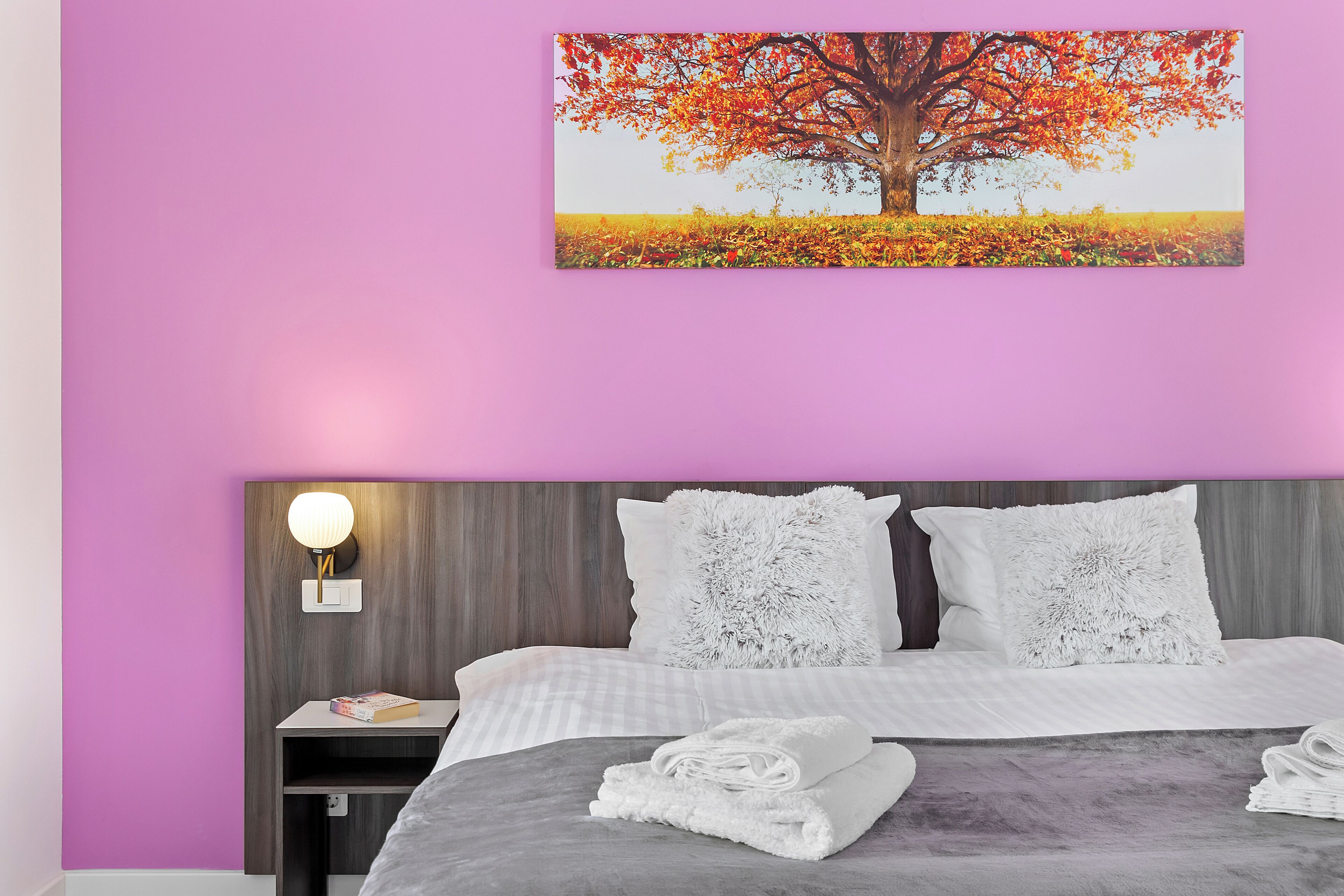 Een slaapkamer met een bed, een nachtkastje, een wandlamp en een schilderij van een boom.