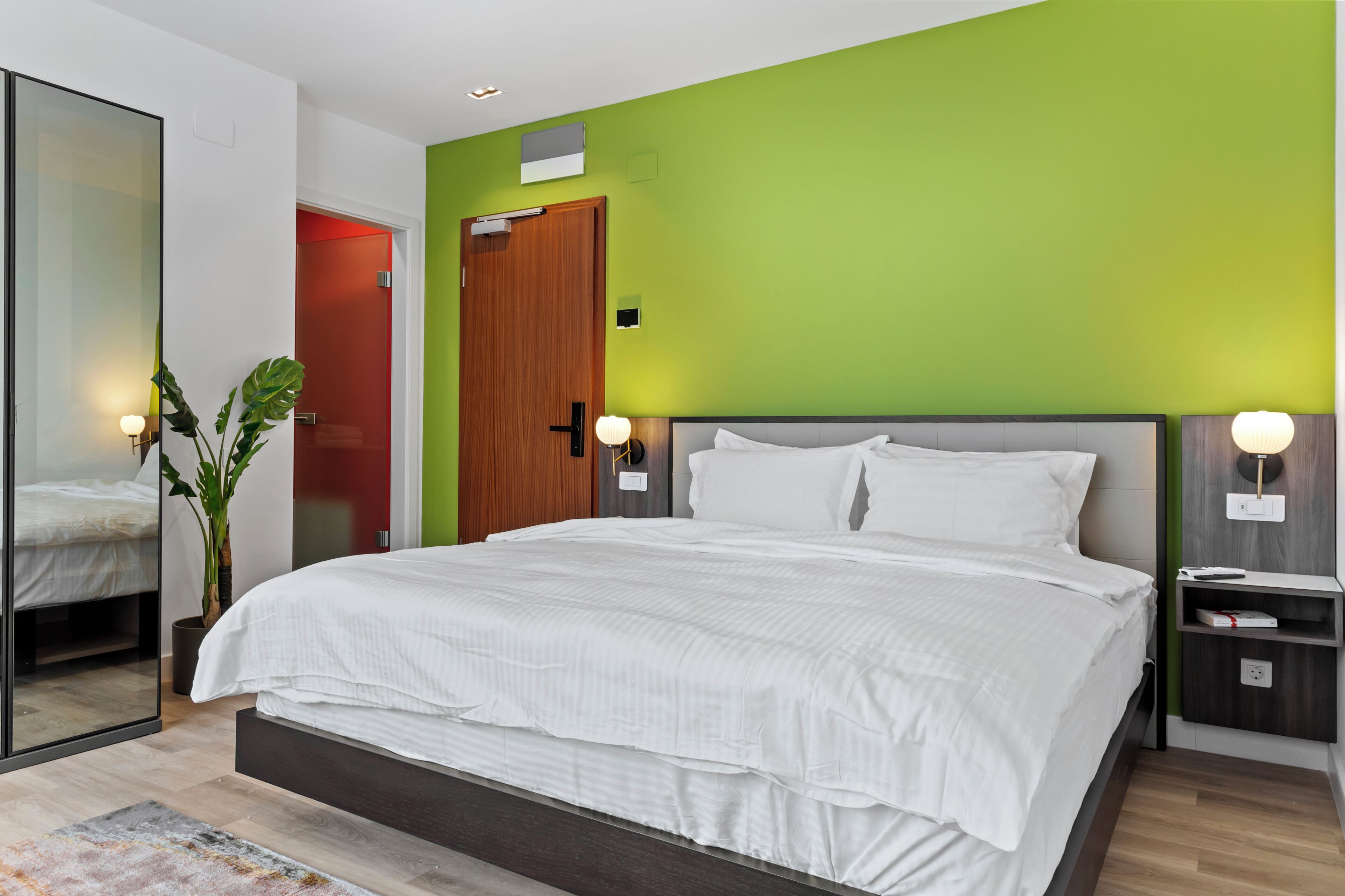 A modern bedroom with a large bed, a bedside table, a nightstand, a mirror, and a green accent wall.