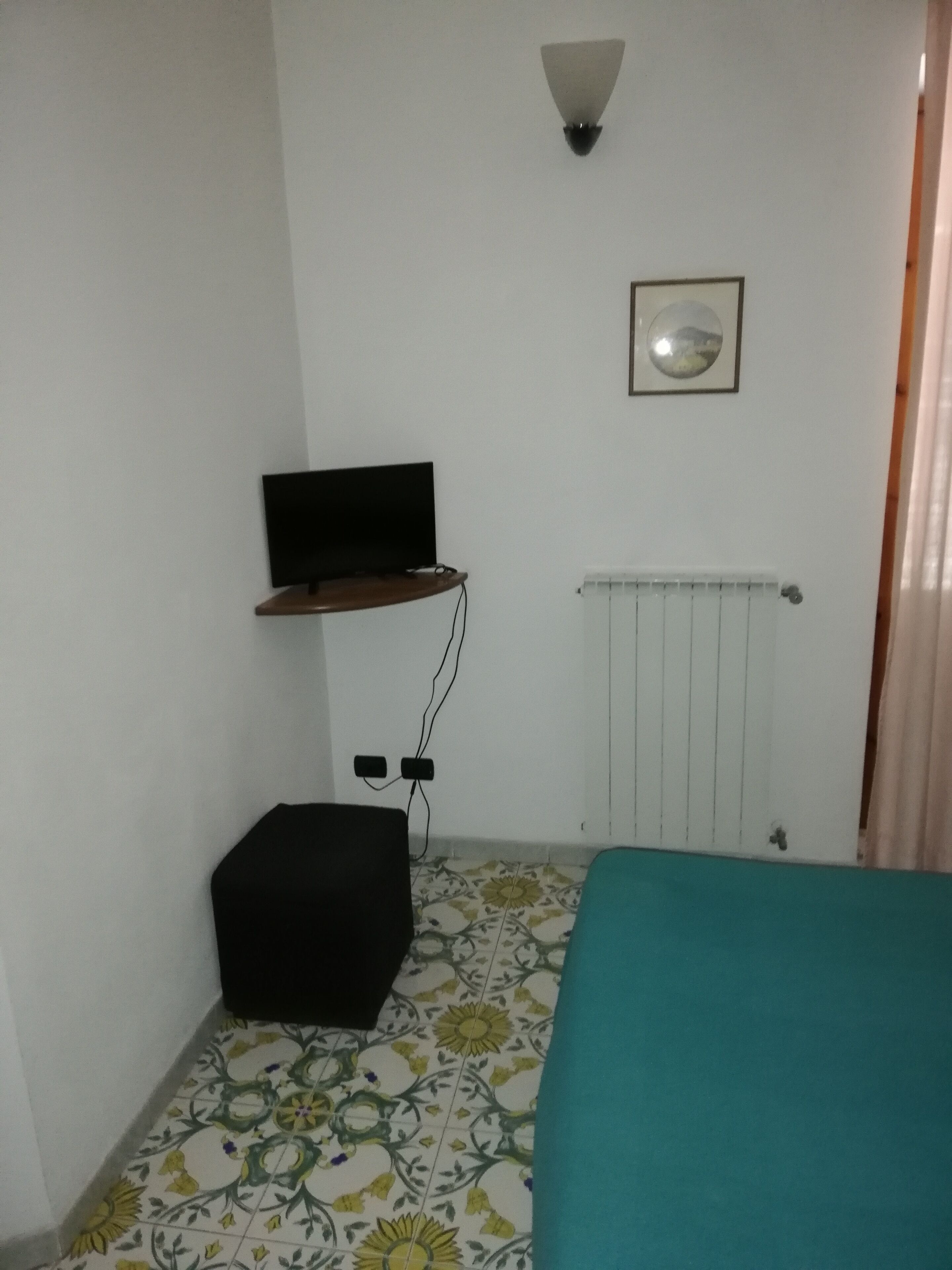 Une petite pièce avec un lit, un téléviseur posé sur une étagère, un radiateur et une prise électrique au mur.