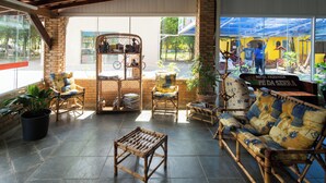 Un espacio interior amplio con un sofá de bambú, una mesa de madera y sillas de mimbre. Hay plantas en macetas y un ventanal con vistas a la calle.