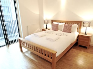 A neatly made bed with white linens and towels, two bedside tables with lamps, and a view of the outside through a sliding door. - Best place to stay and relax patient good to stay  (Teaneck)