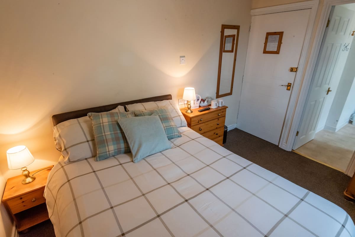 A neatly made bed with a plaid bedspread, a bedside lamp, a wooden nightstand, and a dresser with a mirror.