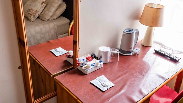 Habitación de hotel con un escritorio de madera, una lámpara, un hervidor, un control remoto y una cama con almohadas.