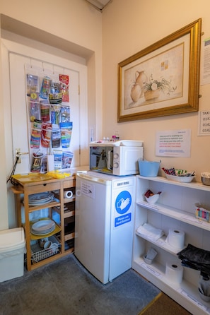 A compact kitchen area with a microwave, a small refrigerator, and a sink. There is a shelf with various items and a framed picture on the wall. - Aarondale Guest House (Hartlepool)
