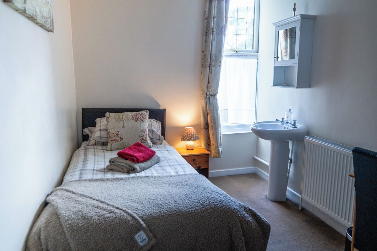 A bedroom with a bed, a bedside lamp, a window with curtains, a radiator, and a small cabinet above the sink.