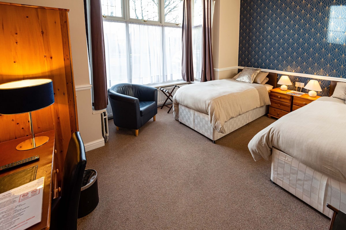 A hotel room with two beds, a chair, a desk with a lamp, and a window with curtains.