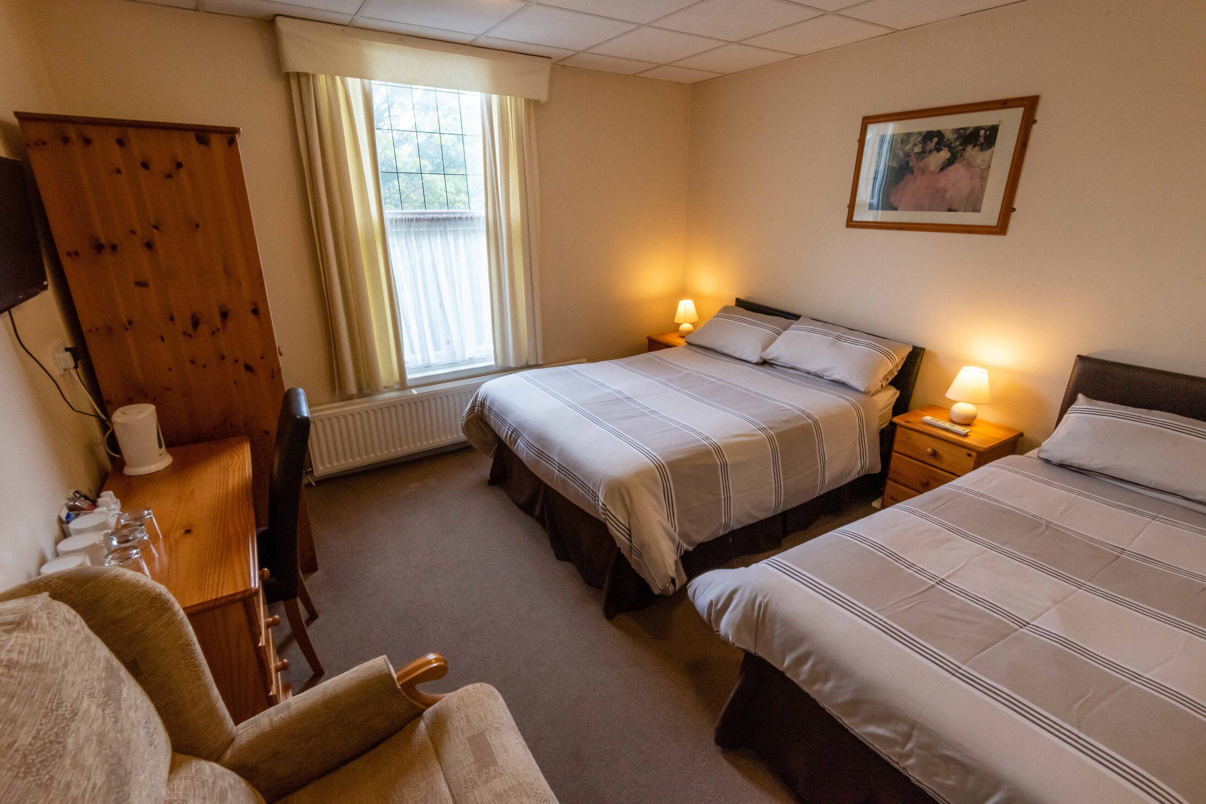 Ein Hotelzimmer mit zwei Betten, einem Schreibtisch mit Stuhl, einem Heizkörper, einem Fenster mit Vorhängen und einem Bild an der Wand.