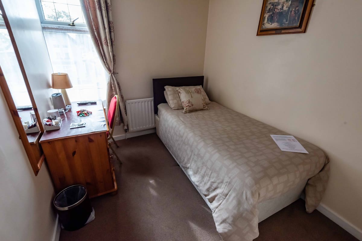 A bedroom with a bed, a wooden desk, a lamp, a radiator, and a window with curtains.