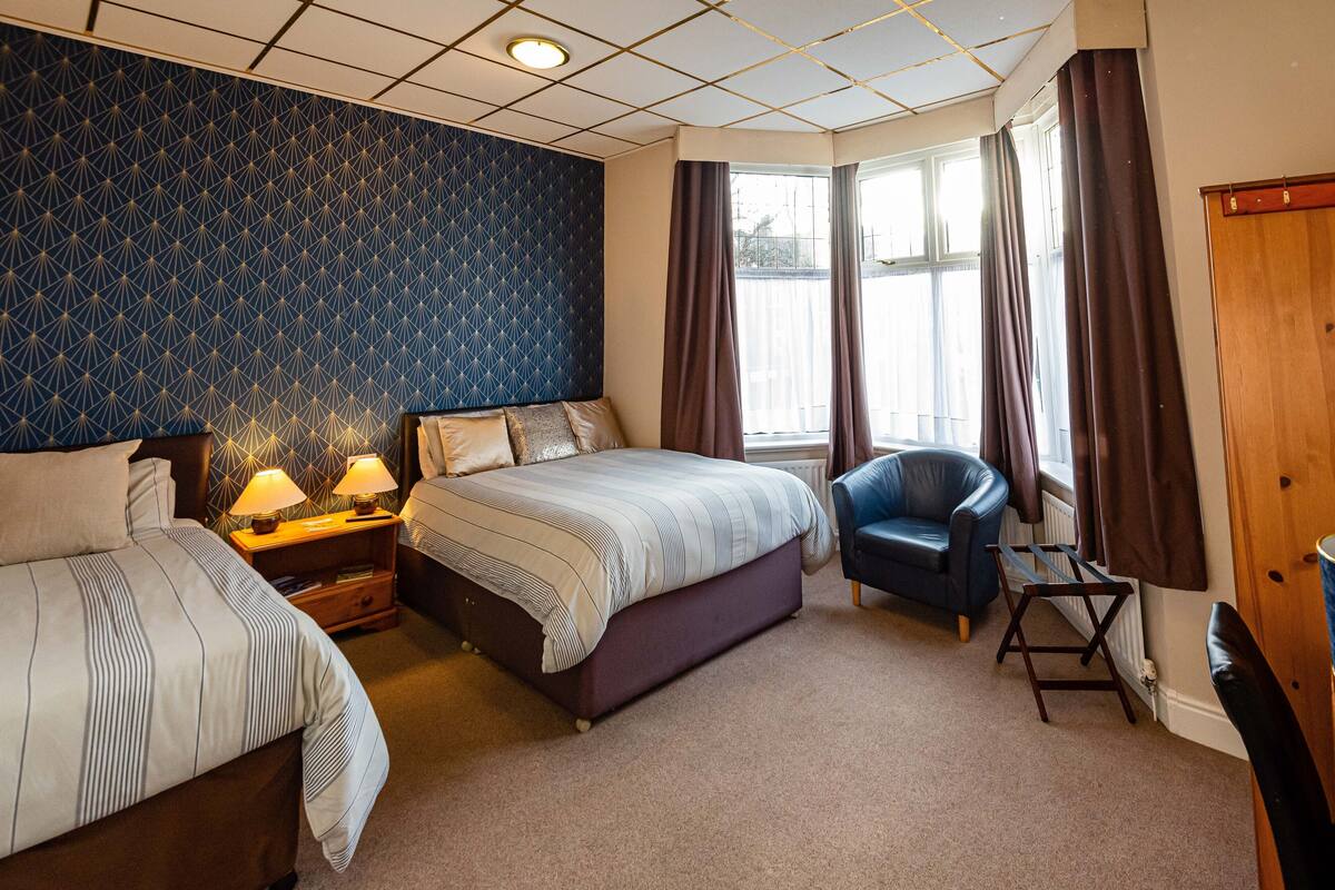 A hotel room with a large bed, two bedside tables with lamps, a blue armchair, and a window with curtains.