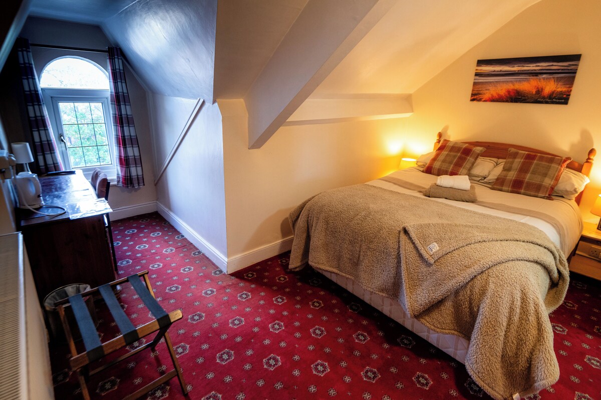 A bedroom with a bed, a window with curtains, a painting on the wall, and a red patterned carpet.