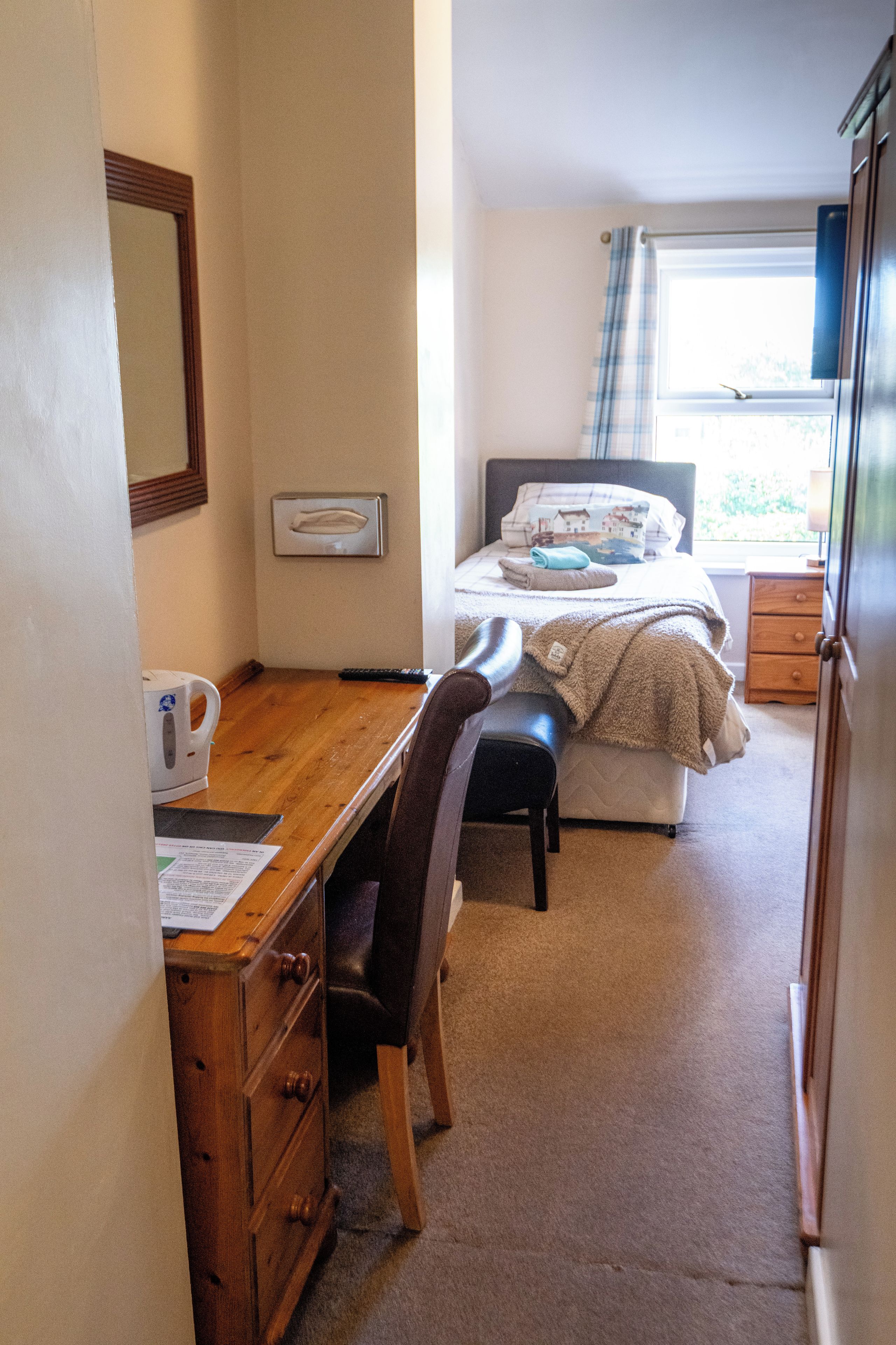 Habitación de hotel pequeña con una cama individual, un escritorio, una silla, una mesita y un ventanal con cortinas.