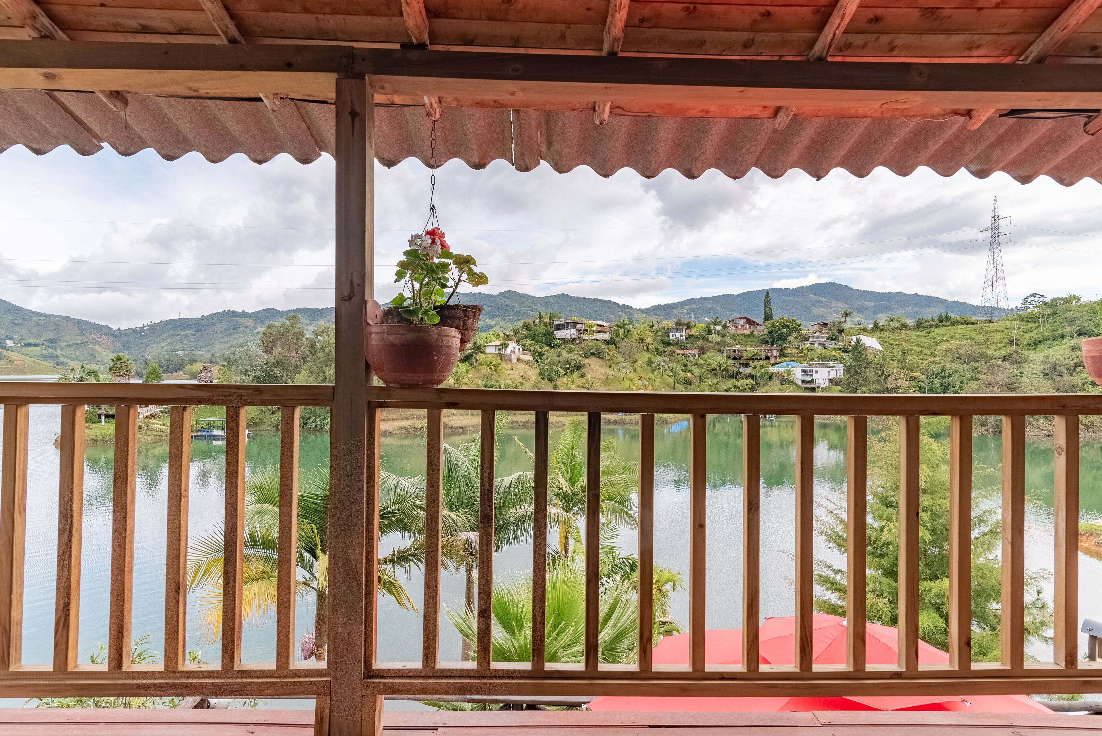 Un balcón de madera con vista a un lago, palmeras y casas a lo lejos.