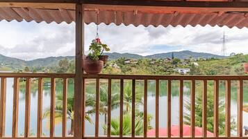 Un balcón de madera con vista a un lago, palmeras y casas a lo lejos.
